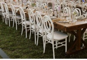 Ornate, white chairs set up agains a wooden table on the grass for a large dinner.