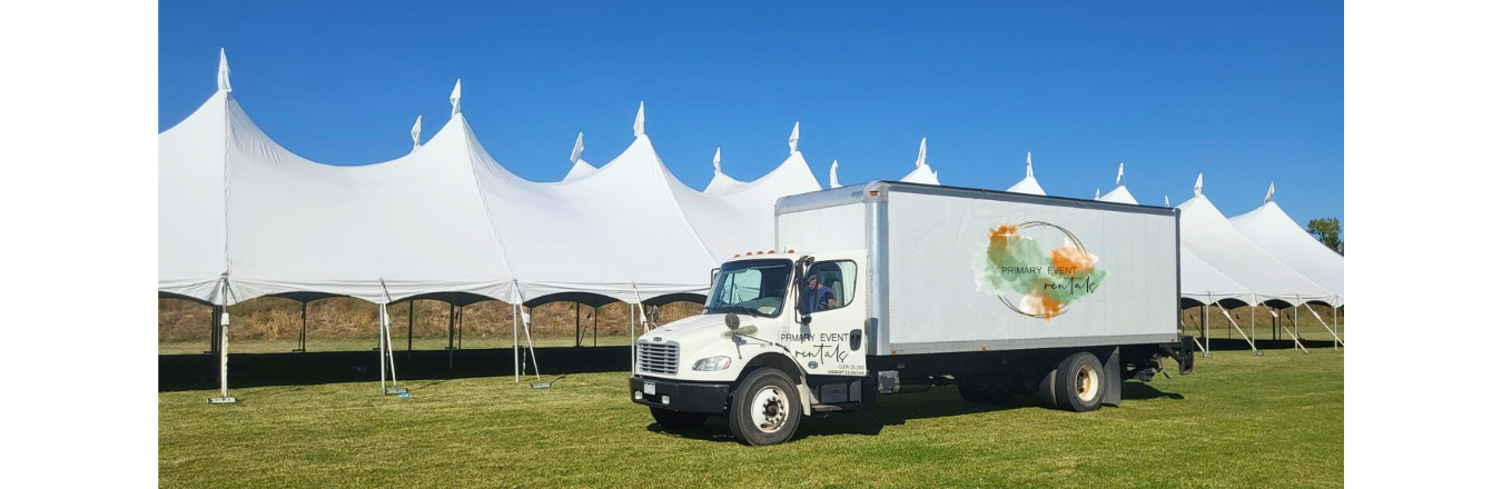 A large delivery truck with the Primary Event Rentals logo is parked on green grass in from of a very large, white, party tent.