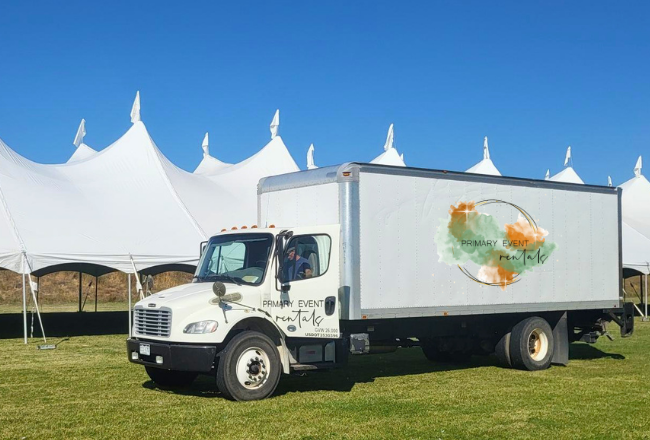 A large delivery truck with the Primary Event Rentals logo is parked on green grass in from of a very large, white, party tent.
