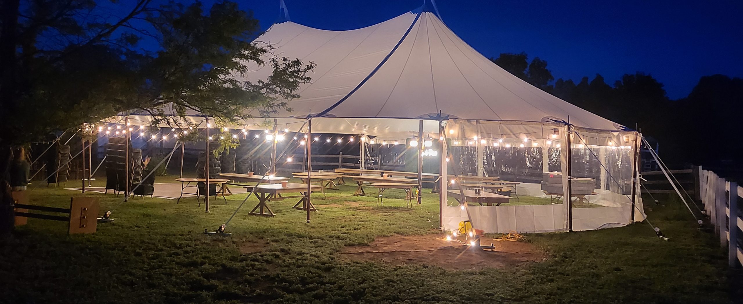 Large white outdoor event tent illuminated with string lights at dusk, featuring picnic tables and clear sidewalls on a grassy lawn.