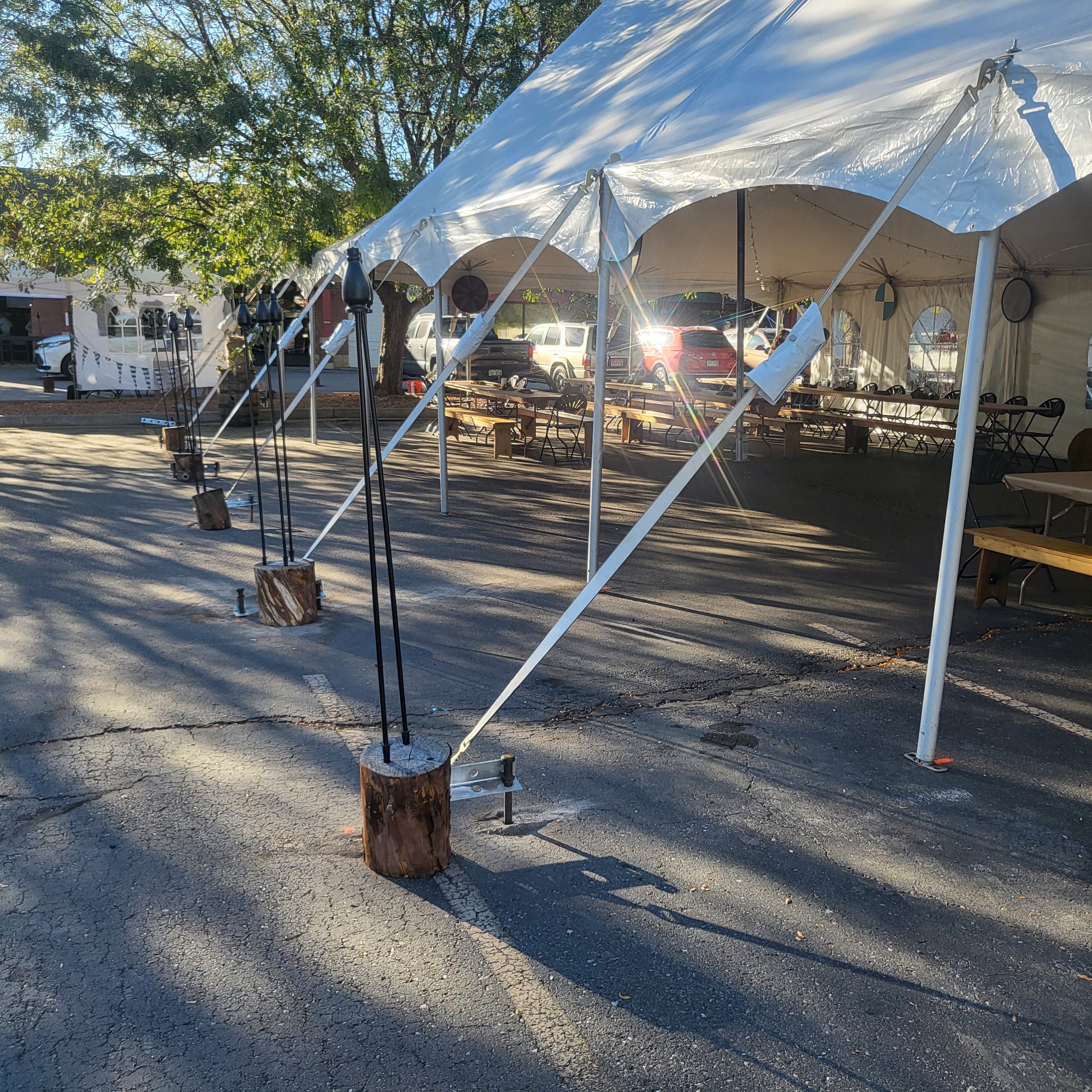 Large white event tent set up over an outdoor paved area with tables and chairs underneath, secured with support straps and rustic wooden weights.
