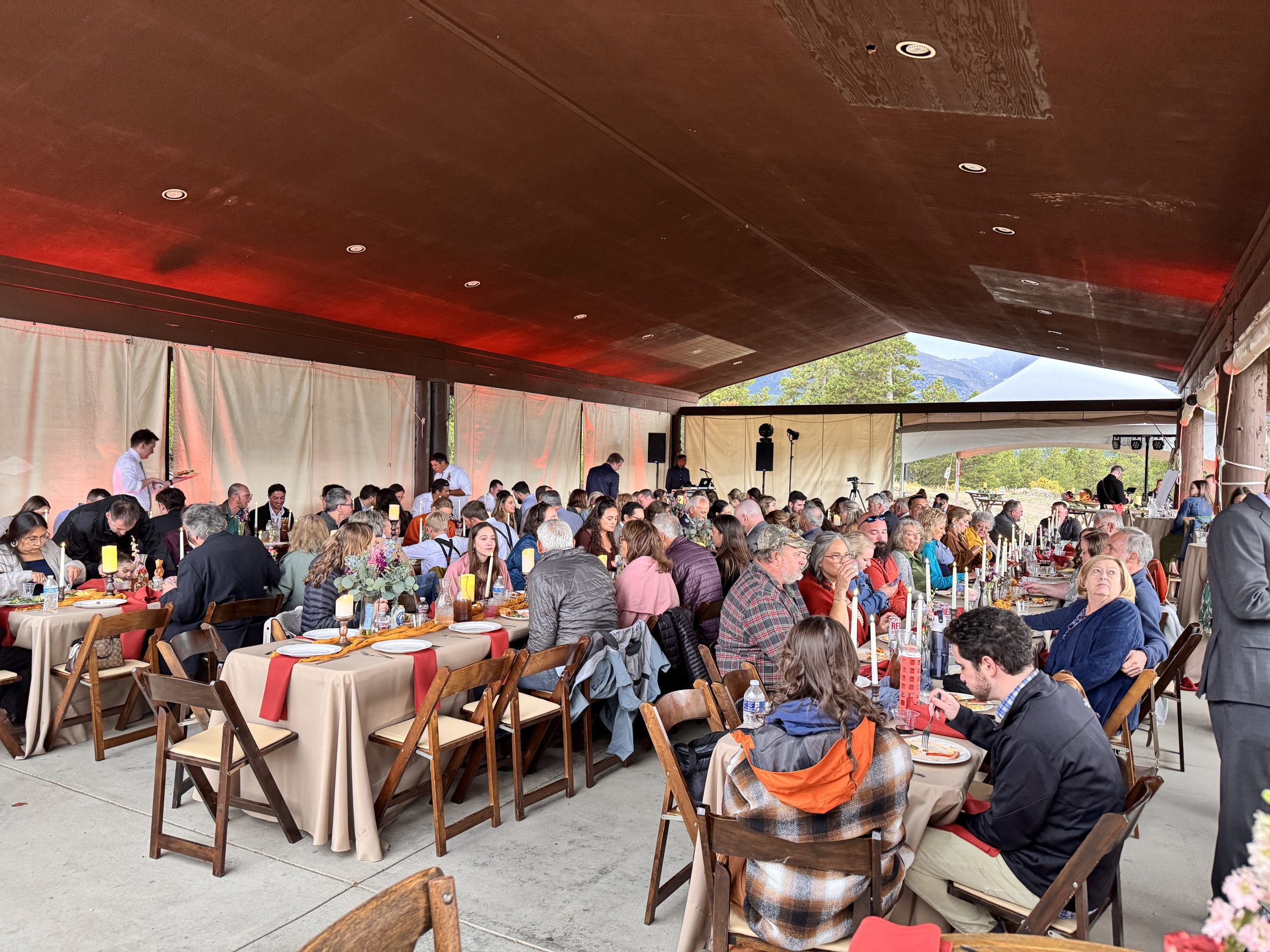 Large outdoor event under a covered pavilion with guests seated at long banquet tables, wooden chairs, neutral linens, red accents, floral centerpieces, and warm event lighting, with mountain views in the background.